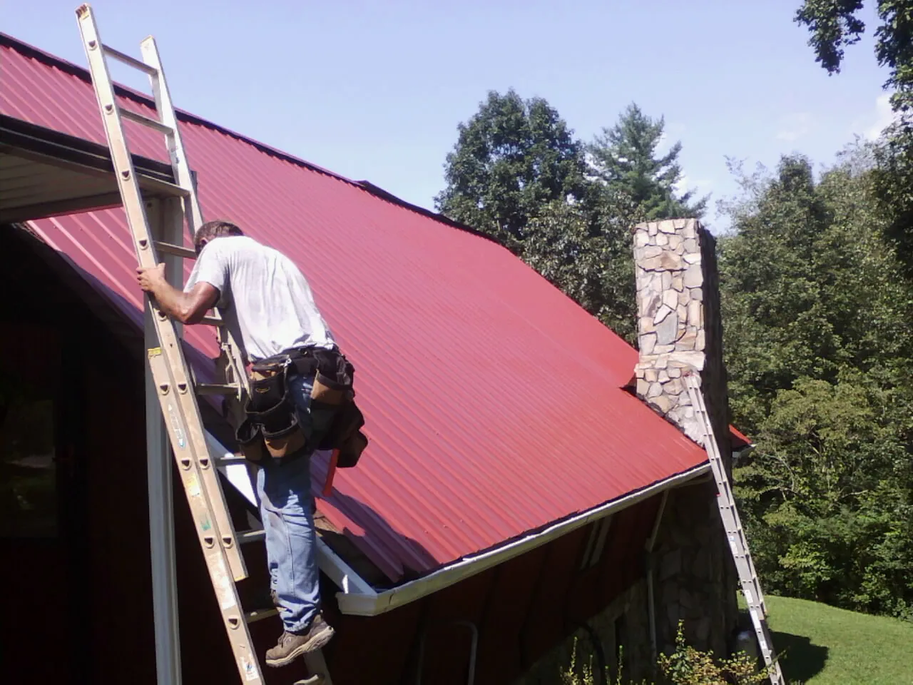 Roofer on a ladder beside a red metal roof and stone chimney — Soffit & Fascia Repair by Roofing Brothers in Montgomeryville